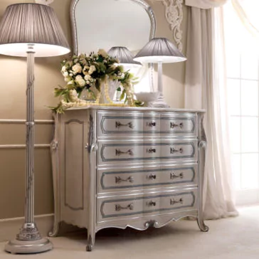 A vintage-style Ornate Italian Chest of Drawers with intricate details features four drawers adorned with decorative handles. Two silver lamps with gray lampshades and a vase of white flowers sit on top. A matching mirror hangs above the Ornate Italian Chest of Drawers. Cream-colored curtains frame a window to the right.