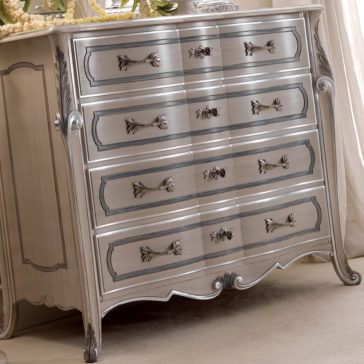 The Ornate Italian Chest of Drawers is a vintage-style, metallic silver dresser featuring five drawers with exquisite metal handles. It boasts detailed carvings on the edges and legs, enhancing its elegance. This refined piece stands on a light-colored carpet.