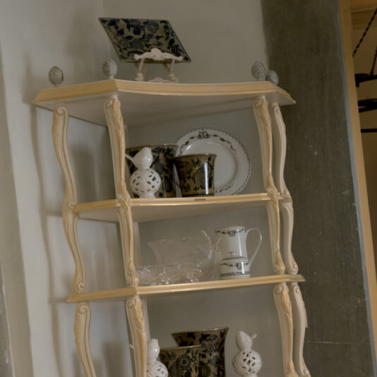 Ornate Italian Designer Corner Shelf An Ornate Italian Designer Corner Shelf, in a decorative cream-colored wooden finish with four levels, displays various items including ceramic vases, decorative plates, cups, and a small teapot against a white wall. This piece features intricate carved details along its edges.