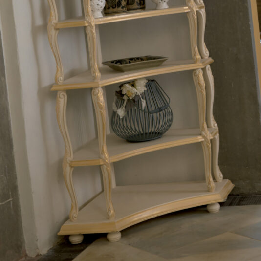 Ornate Italian Designer Corner Shelf Against a wall stands an Ornate Italian Designer Corner Shelf, featuring curved legs and four tiers. The second tier is adorned with a decorative wire vase filled with white flowers, while the third tier supports a small dish. Beneath the shelf, the floor showcases a pattern of light and dark diagonal tiles.