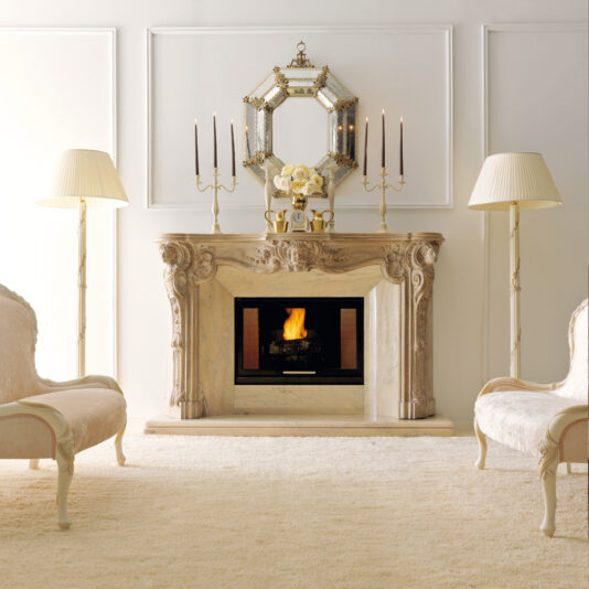 Elegant living room featuring the Reproduction Italian Fireplace Surround with a roaring fire. Above the mantel is a round mirror surrounded by candle holders and a floral arrangement. Two matching floor lamps and beige upholstered chairs flank the fireplace, set against a white wall.