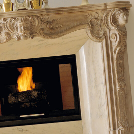 A close-up view of a Reproduction Italian Fireplace Surround showcases its ornate craftsmanship with intricate carvings of faces and scrollwork. A bright fire burns within, casting a warm glow throughout the room. The mantle above the fireplace is adorned with various small golden ornaments, completing the elegant look.