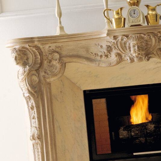 A close-up view of the Reproduction Italian Fireplace Surround showcases its ornate marble structure, embellished with intricate carvings of faces and floral motifs. The elegant display is complemented by two candlesticks and a small clock placed at the center, with a golden pitcher adorning the top. Inside, a warm fire burns brightly.