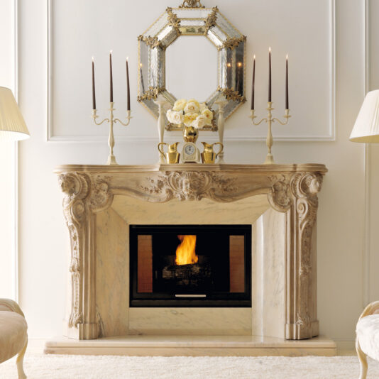 Elegant room with a luxurious Reproduction Italian Fireplace Surround featuring intricate marble carvings. Above the fireplace, there's a decorative octagonal mirror and a golden candelabrum holding white roses. Two gold candleholders with long, dark candles flank the mirror.
