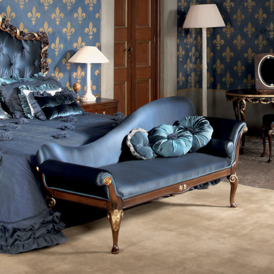 Elegantly decorated bedroom featuring a luxurious Reproduction Two Tone Walnut Italian Chaise Longue adorned with plush pillows in front of an ornate bed. The room has fleur-de-lis patterned wallpaper, a wooden door, bedside tables with lamps, a vintage-style mirror, and a floor lamp.