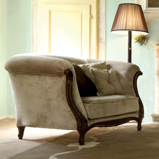 A plush, beige velvet loveseat featuring a classic design with carved wooden legs and arms. It is adorned with three matching cushions. Next to the loveseat, a tall, pleated lampshade on a slender lamp base casts a warm light, perfectly complementing the cozy elegance of the nearby Two Tone Walnut Designer Italian Armchair.