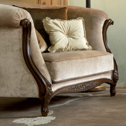 Two Tone Walnut Designer Italian Armchair The Two Tone Walnut Designer Italian Armchair is an elegant vintage piece with intricately carved wooden armrests and legs, upholstered in light beige velvet fabric. It includes a matching square pillow adorned with a floral design and fringed edges on the seat. The chair is set on a beige and cream patterned rug.