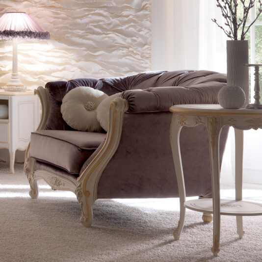 A cozy living room showcases the elegant Venetian Button Upholstered Italian Armchair with its plush cushions, complemented by a round side table adorned with decorative items, a lamp perched on a small wooden table, and textured wall art. Soft, natural light filters through sheer white curtains, creating a warm and inviting atmosphere.
