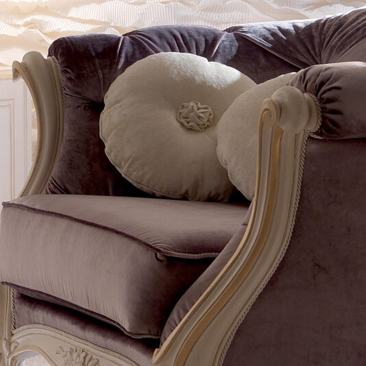 The Venetian Button Upholstered Italian Armchair, a luxuriously upholstered piece in plush velvet with beige trim and two round beige accent pillows, sits in a softly lit room. This chair boasts an ornate wooden frame with intricate carved details and exudes rich sophistication.