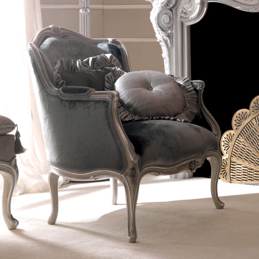 A luxurious Venetian Designer Italian Armchair, featuring plush grey fabric and ornate wooden detailing, sits next to an elegant yet empty bookshelf. The vintage-style armchair is adorned with a button-tufted seat and decorative throw pillows. Soft, natural light brightens the setting.