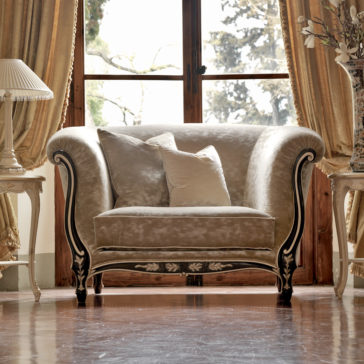 Venetian Designer Italian Armchair A luxurious room featuring a plush, ornate Venetian Designer Italian Armchair with curved armrests, two accent pillows, tall windows framed by elegant drapes, and a floor lamp and side table with a potted plant on either side of the chair, all on a polished wooden floor.