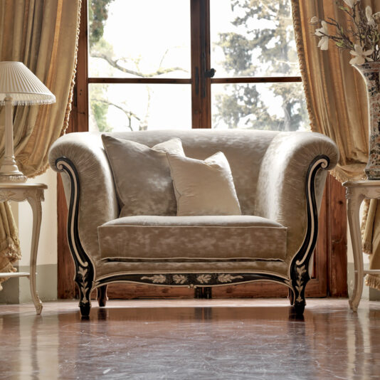 A luxurious room featuring a plush, ornate Venetian Designer Italian Armchair with curved armrests, two accent pillows, tall windows framed by elegant drapes, and a floor lamp and side table with a potted plant on either side of the chair, all on a polished wooden floor.
