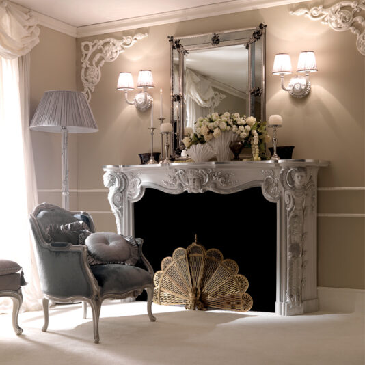 The opulent living room features an ornate white fireplace adorned with a floral arrangement and symmetrical wall sconces. A large decorative fan screen covers the fireplace opening. The luxurious, classical decor is complemented by a Venetian Designer Italian Armchair, a matching floor lamp, and an elegant grey upholstered chair.