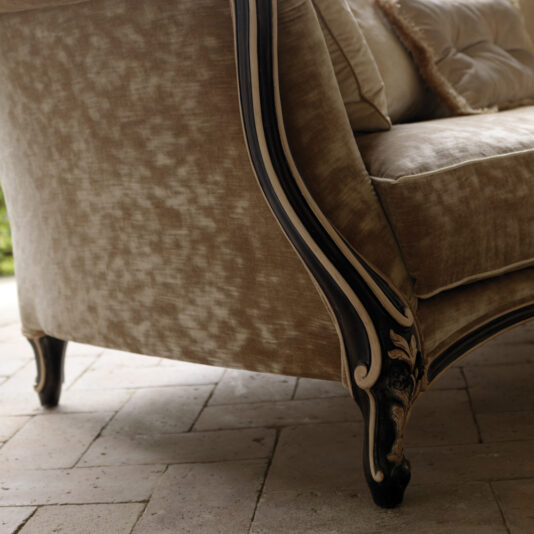 Venetian Designer Italian Armchair A close-up angle of an elegant Venetian Designer Italian Armchair with ornate, dark wooden legs and plush, light brown fabric. The armchair is placed on a tiled floor, with a soft cushion visible on the seat. The background is blurred, focusing attention on the intricate details of this designer piece.