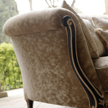 Venetian Designer Italian Armchair Close-up of a plush, beige sofa with ornate, dark wood detailing on the armrest. Just beside it sits an elegant Venetian Designer Italian Armchair. The background shows a sunlit outdoor area with green foliage and a wrought iron railing, giving a sense of a tranquil garden setting.