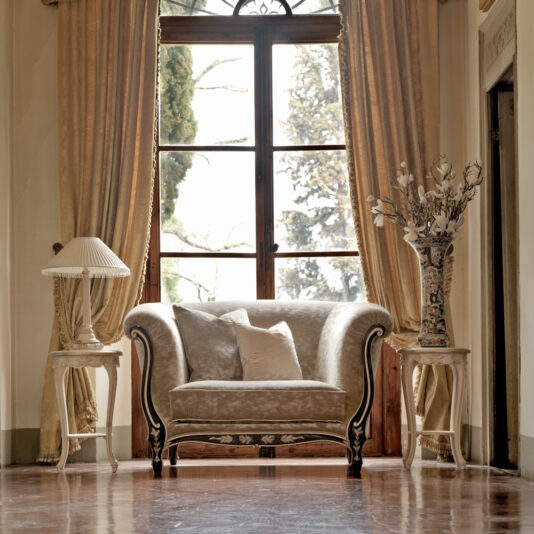 Venetian Designer Italian Armchair An elegant living room showcases a plush Venetian Designer Italian Armchair in beige, complete with two cushions, positioned between two side tables. One table supports a contemporary lamp, while the other presents a decorative vase filled with flowers. Large windows adorned with golden drapes offer a serene outdoor view.