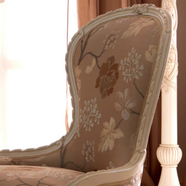 Venetian Style Italian Armchair The Venetian Style Italian Armchair is a vintage piece with a high backrest, showcasing a floral pattern in shades of beige and brown. It features a curved, ornate wooden frame in light cream and is placed near a window adorned with light brown curtains.