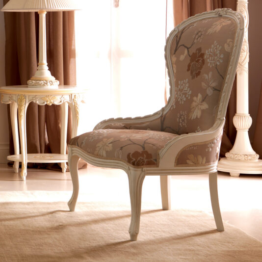 A Venetian Style Italian Armchair featuring an ornate wooden frame and floral fabric upholstery sits on a beige rug. The chair is positioned in a softly lit room with earth-tone drapes, an elegant side table, and a tall decorative floor lamp in the background.