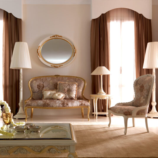 Elegant living room with a classic style sofa, a Venetian Style Italian Armchair, and a round mirror on the wall. The furniture has intricate golden detailing and floral upholstery. Two tall lamps stand beside the sofa and armchair. The room features large windows with brown curtains.