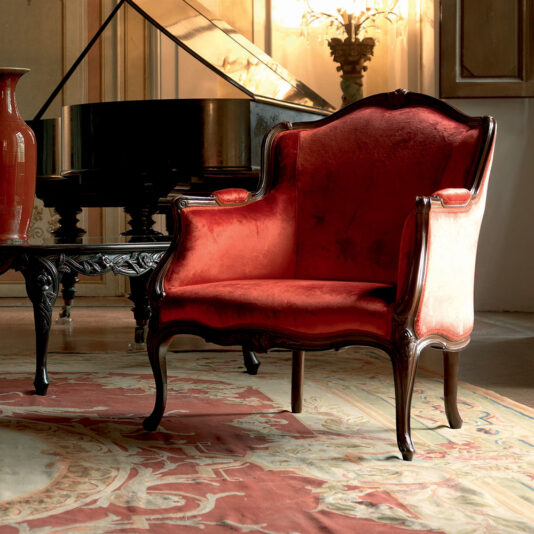 A Venetian-style room features a Venetian Style Walnut Italian Armchair adorned in luxurious red velvet, complete with ornate wooden legs and armrests. It sits elegantly on a patterned rug in front of a grand piano, accompanied by a decorative table and a large red vase. Warm, ambient lighting enhances the classic ambiance of the space.