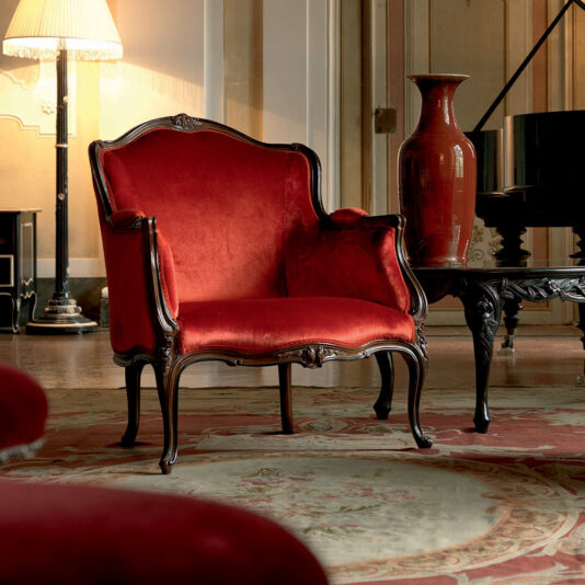A luxurious room features a Venetian Style Walnut Italian Armchair adorned with rich red velvet, placed upon an elegant rug. Nearby, a large red vase sits on a small table, and in the background, a lamp and piano enhance the sophisticated ambiance.
