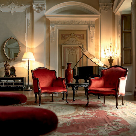 A luxurious, ornate room featuring two Venetian Style Walnut Italian Armchairs upholstered in red in the foreground, a grand piano in the corner, and elegant decor including an ornate rug, statues, a table with a red vase, a large mirror, and an ornate lamp. The room has intricate wall moldings and warm lighting.
