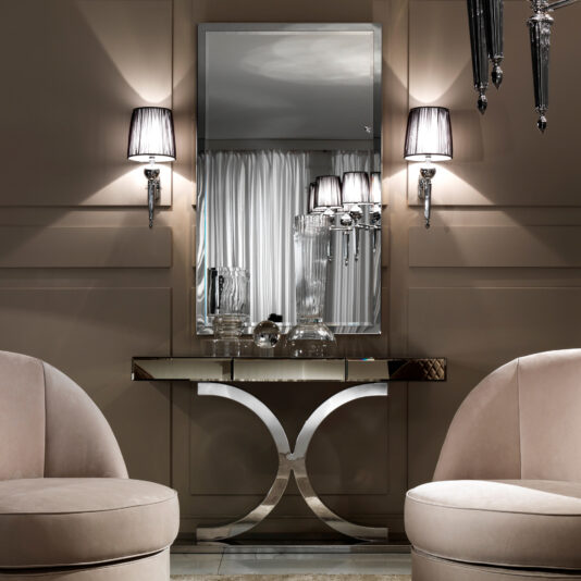 A sophisticated atmosphere is created with a modern console table beneath a Large Gold Italian Wall Mirror, flanked by two warm-glow wall-mounted lamps. Two plush, rounded armchairs face each other elegantly.