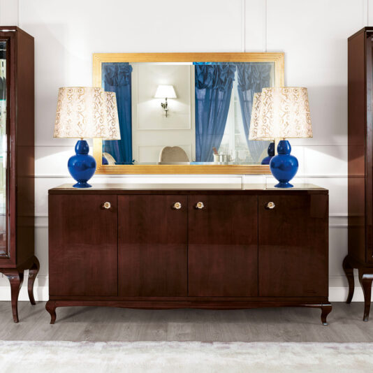 High end Modern Glamour large Wenge Sideboard A High end Modern Glamour large Wenge Sideboard with a polished finish and ornate brass handles stands in front of a large mirror framed in gold. Two decorative blue lamps with patterned shades are symmetrically placed on the sideboard. Blue curtains reflect in the mirror, adding a touch of modern glamour.