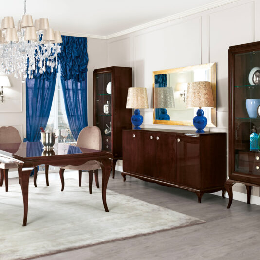 High end Modern Glamour large Wenge Sideboard A luxurious dining room features a dark wooden table surrounded by beige upholstered chairs. A chandelier hangs above, and a large blue curtain adorns the window. The High-End Modern Glamour Large Wenge Sideboard, complemented by blue lamps and a large mirror and flanked by glass cabinets, adds high-end modern glamour to the setting.