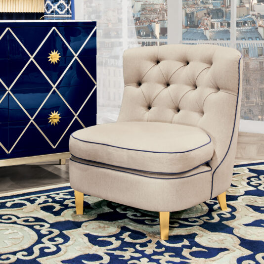 A High End Modern Italian Occasional Armchair with beige tufted upholstery and gold legs sits on a patterned blue and white rug. Behind this chair is a dark blue dresser adorned with a gold starburst pattern and diamond design. In the background, a cityscape with buildings and windows is visible.