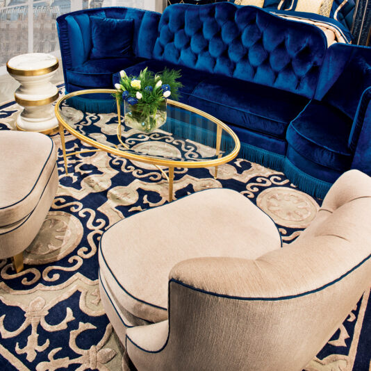 A luxurious living room with a blue velvet sofa, a round glass coffee table with gold trim, High End Modern Italian Occasional Armchairs in beige, and a floral-patterned rug. A bouquet of flowers sits on the table, and the room is tastefully decorated with elegant furnishings.