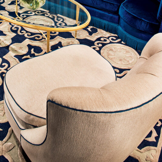 A High End Modern Italian Occasional Armchair with beige upholstery and a matching ottoman sits on an ornate navy and gold patterned rug. A gold-framed glass coffee table is partially visible next to the chair. The background features a navy blue, cushioned wall or furniture element.
