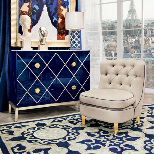 A stylish living room setup features a deep blue dresser adorned with gold star designs, a marble bust, a lamp, and artwork. A High End Modern Italian Occasional Armchair sits on a patterned blue and white rug beside the beige tufted chair. Large windows reveal a cityscape with the Eiffel Tower in the background.