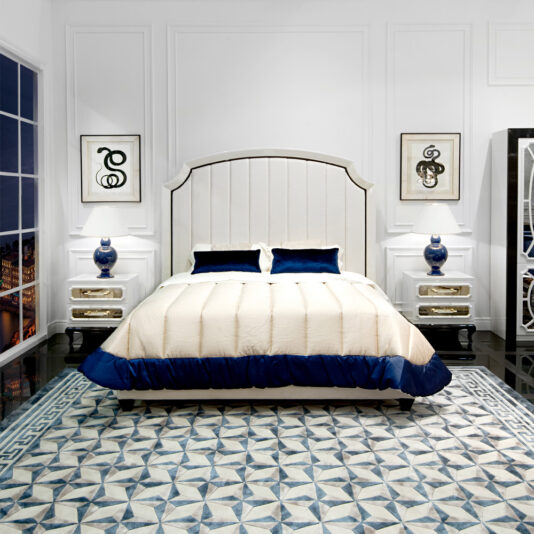 A high-end modern bedroom featuring the High End Modern White Ottoman Storage Bed adorned with neatly made white and blue bedding. The room boasts a geometric-patterned rug, two bedside tables with blue lamps, framed artwork, and a large window offering breathtaking cityscape views.