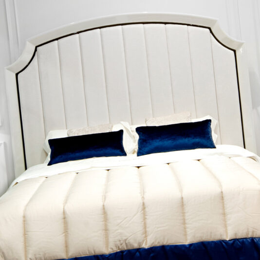 The High End Modern White Ottoman Storage Bed boasts a luxurious cream-colored design, enhanced by a tall, curved headboard. The bed is dressed in neatly arranged cream-colored bedding, complemented by two dark blue velvet pillows and a dark blue throw at the foot. White paneled walls serve as an elegant backdrop.