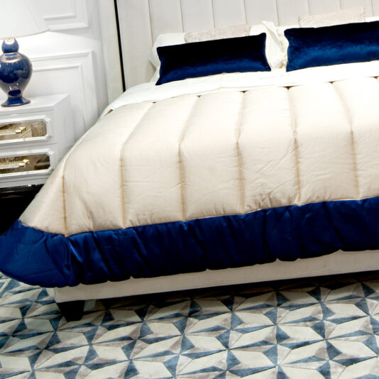 A neatly made High End Modern White Ottoman Storage Bed with a white and blue comforter, matching blue pillows, and a cream upholstered headboard. A white nightstand with drawers and a blue lamp sits on the left. The floor features a geometric pattern in shades of blue and white.