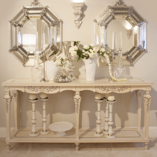 The Classic Italian Large Console Table with drawers is a stylish and timeless piece, beautifully adorned with white flowers in a vase, crystal candle holders, and a silver bowl. Above the table hang two ornate mirrors that reflect the room. This large cream-colored console table features intricate carvings and convenient drawers, complemented by standing candlesticks below.