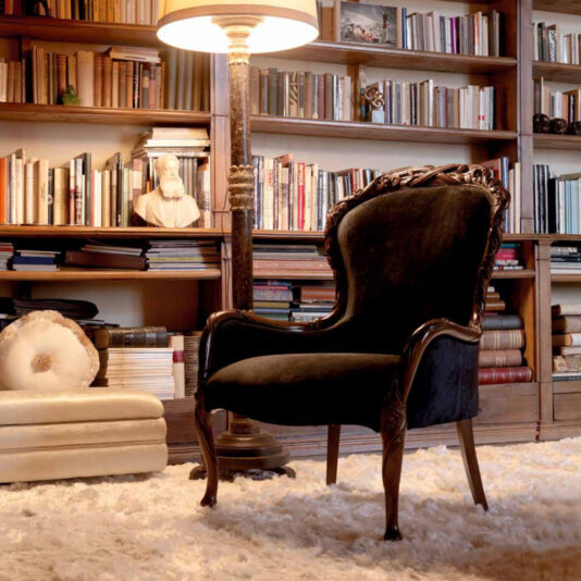 A cozy reading nook featuring a dark upholstered armchair with ornate wood carvings, placed on a plush white rug. A tall floor lamp illuminates the space, which is flanked by bookshelves filled with various books and decor, including a bust statue and a Classic Italian Velvet Rose And Ribbon Walnut Armchair.