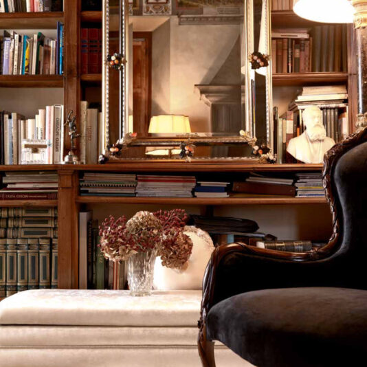 A cozy library with wooden bookshelves filled with various books. A large ornate mirror hangs above a shelf, reflecting a lamp. A white bust of a bearded figure sits beside the mirror. A vase of dried flowers rests on a white ottoman, and a Classic Italian Velvet Rose And Ribbon Walnut Armchair is in the foreground.