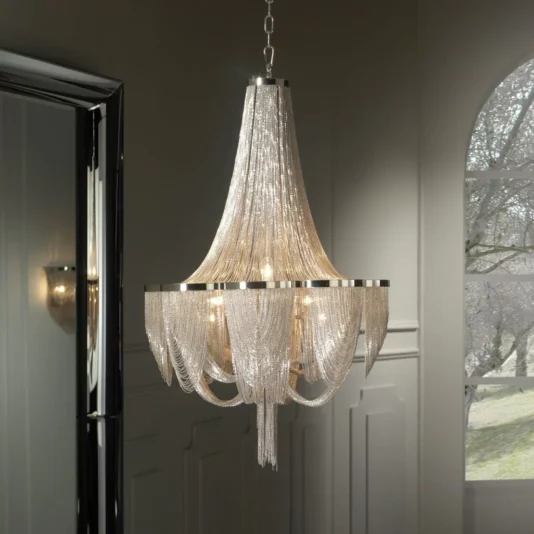 An Empire Silver Chain Chandelier graces the ceiling with its cascading strands of silver chains, perfectly complementing the elegantly decorated room. The space features white walls, a large mirror on one side, and a window that reveals a serene outdoor view with trees.