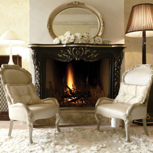 A cozy living room features an elegant fireplace with a gentle fire burning. Two plush Italian Ivory And Silver Rose And Ribbon Armchairs, adorned with ornate detailing, face the fireplace and are set on a luxurious, light-colored rug. Above the fireplace, a decorative oval mirror and a vase of white flowers are displayed.