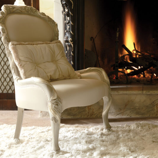 An Italian Ivory And Silver Rose And Ribbon Armchair, featuring plush upholstery and intricate decorative woodwork, is adorned with a cozy pillow and placed on a fluffy white rug. It sits before a roaring fireplace with a black wrought-iron screen, creating a scene that exudes warmth and comfort.