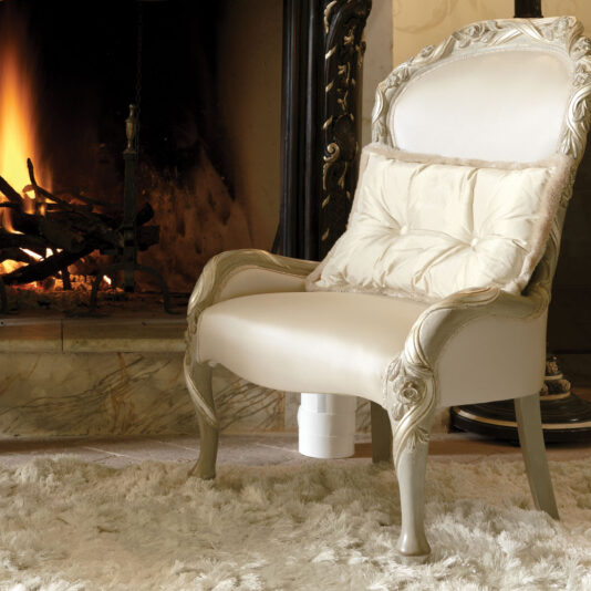 The Italian Ivory And Silver Rose And Ribbon Armchair, adorned with intricate designs, is positioned on a fluffy white rug in front of a warmly lit fireplace. The fire burns brightly, casting a cozy glow on the elegant furniture and creating a luxurious and inviting atmosphere.