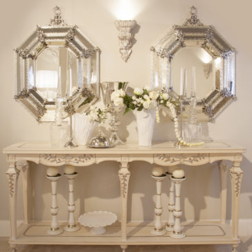 Reproduction Italian Antique Finish Silver Mirror A vintage console table adorned with decorative items, including white candles, flower vases with white flowers, and silver accents, stands against a beige wall. Above the table hang two ornate Reproduction Italian Antique Finish Silver Mirrors in an octagonal shape and a decorative wall sconce.