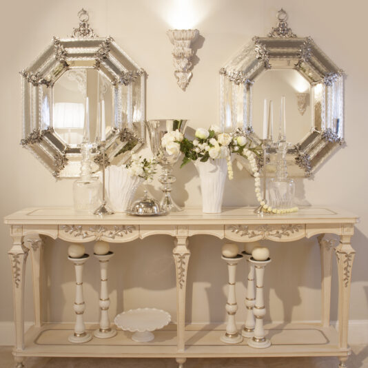 A vintage console table adorned with decorative items, including white candles, flower vases with white flowers, and silver accents, stands against a beige wall. Above the table hang two ornate Reproduction Italian Antique Finish Silver Mirrors in an octagonal shape and a decorative wall sconce.