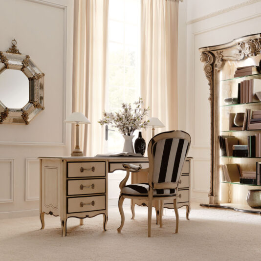 A classic, elegant home office with cream-colored walls and beige curtains. The room features a Reproduction Ivory And Black Italian Desk with multiple drawers, a striped upholstered chair, a decorative mirror, and a bookshelf with ornate detailing. A vase with flowers and table lamps adorn the desk.
