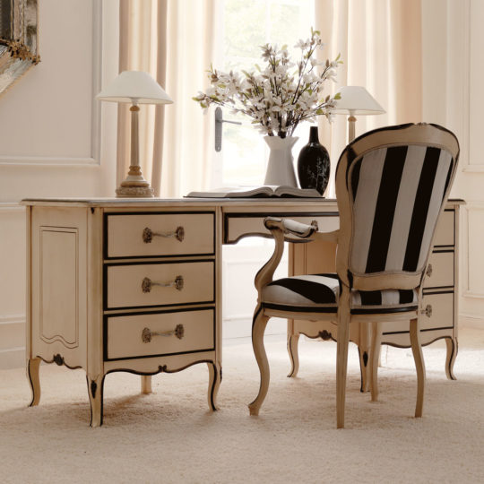 An elegant Reproduction Ivory And Black Italian Desk in a Rococo style featuring four drawers on each side with ornate handles, accompanied by a matching chair adorned with black and white striped upholstery. The desk is complemented by a pair of table lamps and a vase of white flowers. The room is finished with light-colored curtains and carpet.