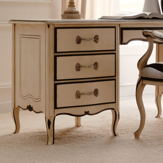 The Reproduction Ivory And Black Italian Desk features three drawers with ornate handles and intricately carved legs that complement its classic design. Partially visible beside the desk is a chair with a cushioned seat, all set up in a well-lit room with carpet flooring.