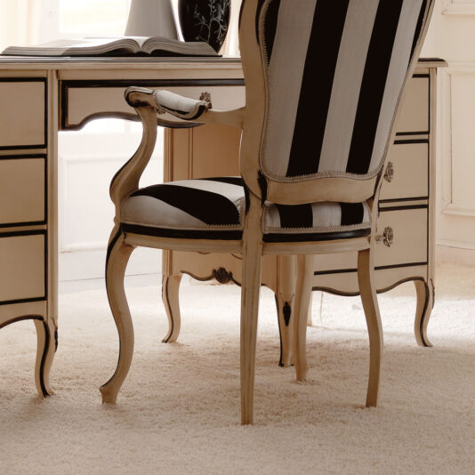 In a well-lit room, set on a light-colored carpet, is the luxurious Reproduction Ivory And Black Italian Desk paired with an elegant chair. The desk showcases ornate detailing and several drawers, and the matching chair boasts a high back with black-and-white striped upholstery. Resting on the desk are an open book and a vase.