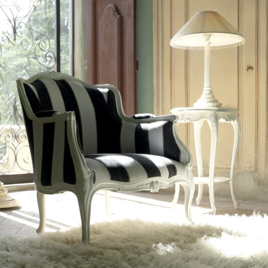 A Venetian Style White Italian Armchair with bold black and white stripes is positioned on a fluffy white rug. Beside it is an elegant side table with a white lamp. Sunlight streams through a window with ornate ironwork, casting a soft glow over the room's classic décor.